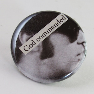 May include: A black and white button with a photo of a person's face and the text "God commanded" printed on a white label.