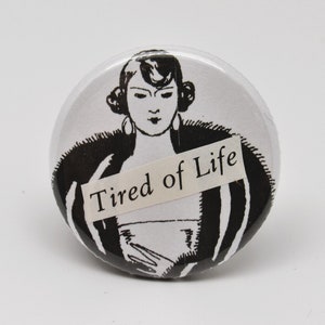 May include: Black and white button featuring a woman with a dramatic expression wearing a fur coat and a white dress. The text "Tired of Life" is printed on a white banner across the image.