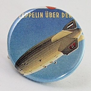 May include: A round button with a blue background and a picture of a zeppelin flying in the sky. The text "Zeppelin über den" is printed on the button.