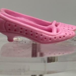 May include: A pair of small, light pink doll shoes. The shoes have a low heel, a rounded toe, and a strap across the top. The shoes are made of a solid material and have small holes throughout the design.