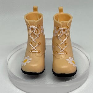 May include: A pair of beige ankle boots with white daisy print and white laces. The boots have a black sole.