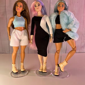 May include: Three fashion dolls with different hairstyles and outfits. The doll on the left is wearing a blue tank top, white shorts, and beige boots. The doll in the middle is wearing a black and white dress and beige boots. The doll on the right is wearing a black crop top, black shorts, a light blue fur coat, and beige boots.