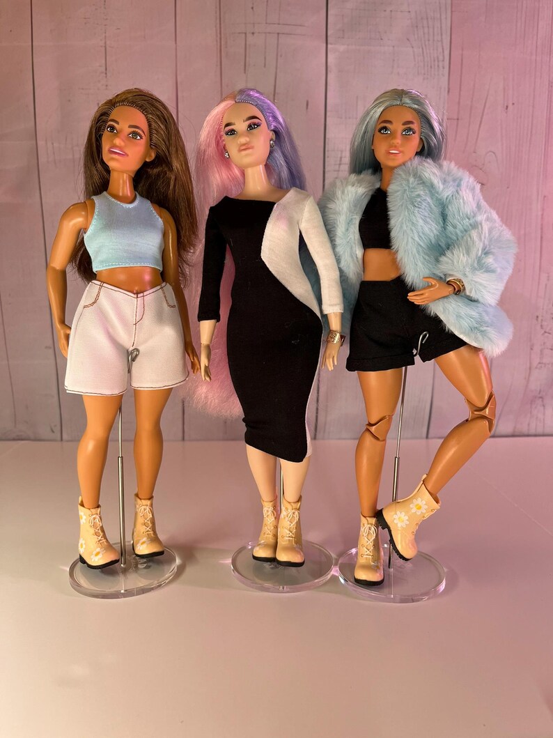 May include: Three fashion dolls with different hairstyles and outfits. The doll on the left is wearing a blue tank top, white shorts, and beige boots. The doll in the middle is wearing a black and white dress and beige boots. The doll on the right is wearing a black crop top, black shorts, a light blue fur coat, and beige boots.