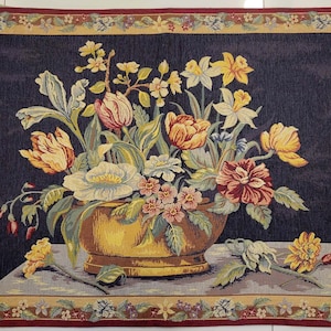 May include: A rectangular woven tapestry featuring a dark blue background with a central golden bowl filled with a colorful floral arrangement of tulips, daffodils, and small blossoms. The piece is framed by a decorative border of flowers.