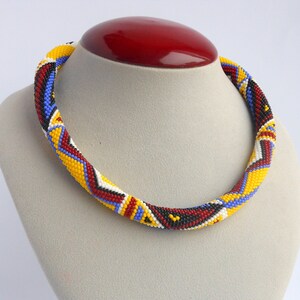 May include: A beaded necklace with a geometric pattern in red, yellow, blue, black, and white. The necklace is displayed on a gray mannequin bust with a red base.