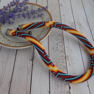 Op de afbeelding: Een kleurrijke kralenketting met een geometrisch patroon in rood, geel, blauw en wit. De ketting ligt op een houten oppervlak met een decoratieve schotel en lavendel takjes.
