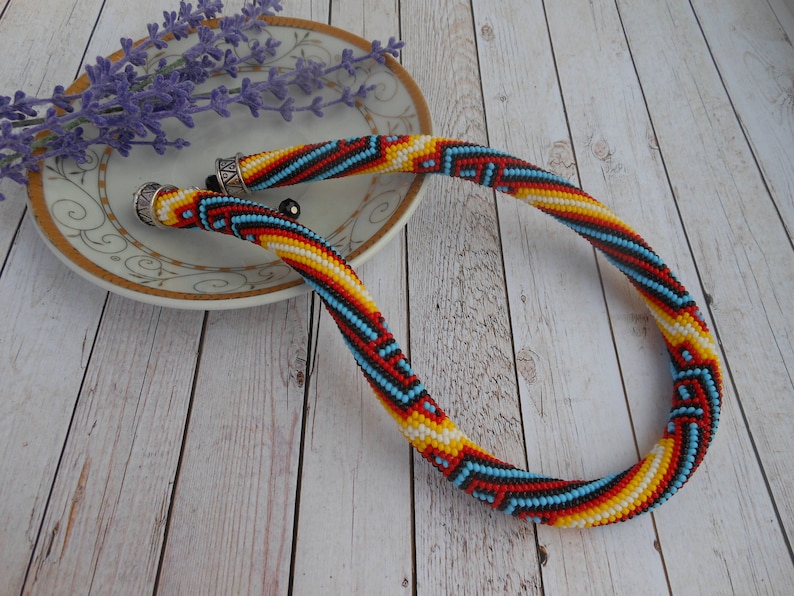 Op de afbeelding: Een kleurrijke kralenketting met een geometrisch patroon in rood, geel, blauw en wit. De ketting ligt op een houten oppervlak met een decoratieve schotel en lavendel takjes.