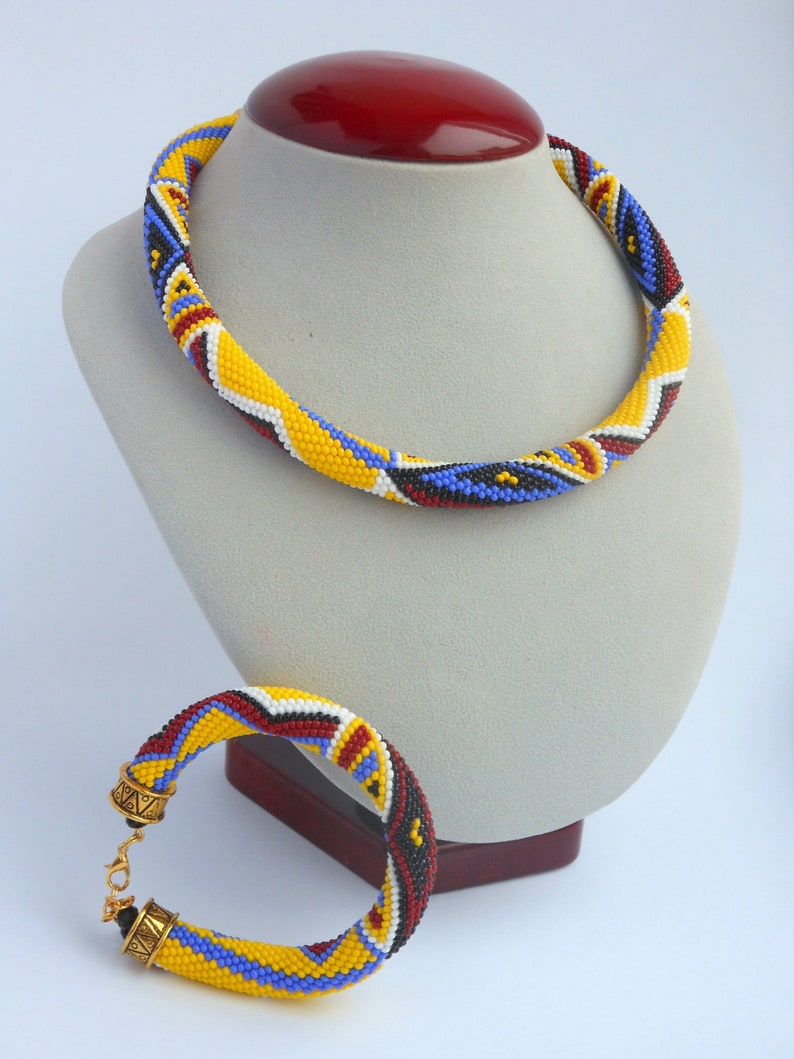 May include: A beaded necklace and bracelet set in yellow, blue, red, white, and black. The necklace is displayed on a mannequin and the bracelet is lying on a table.