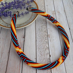 Op de afbeelding: Een kralenketting met een spiraalvormig ontwerp. De ketting heeft een patroon van rode, gele, blauwe en zwarte kralen. De ketting wordt gepresenteerd op een lichtgekleurd houten oppervlak.