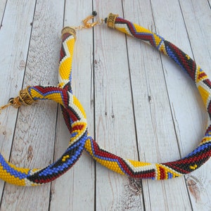 May include: A beaded necklace and matching earrings with a geometric pattern in yellow, red, white, and blue colors. The necklace has a gold clasp and the earrings have gold hooks.