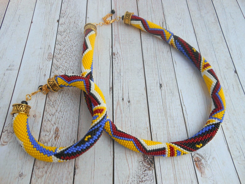 May include: A beaded necklace and matching earrings with a geometric pattern in yellow, red, white, and blue colors. The necklace has a gold clasp and the earrings have gold hooks.