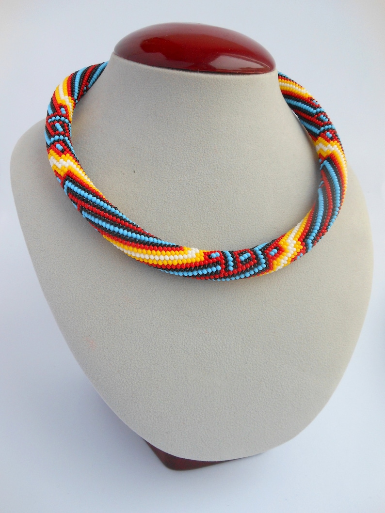 Op de afbeelding: Een kleurrijke kralenketting op een paspop. De ketting heeft een spiraalpatroon met rode, gele, witte, zwarte en blauwe kralen. De kralen zijn dicht op elkaar geregen, waardoor een textuur ontstaat. Een levendig accessoire.