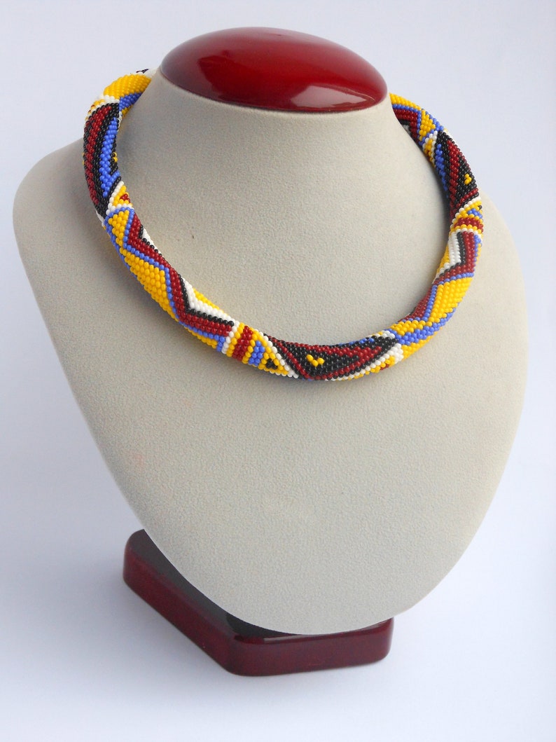 May include: A beaded necklace with a geometric pattern in red, yellow, blue, black, and white. The necklace is displayed on a gray mannequin bust with a red base.