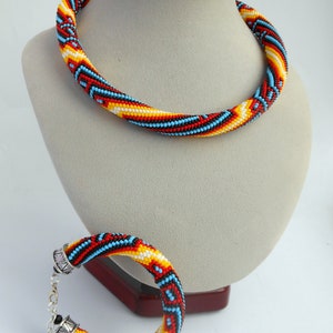 Op de afbeelding: Een kralenketting en armband set. De sieraden hebben een spiraalvormig ontwerp met rode, gele, blauwe en zwarte kralen. De ketting wordt op een mannequin getoond en de armband ligt ernaast.