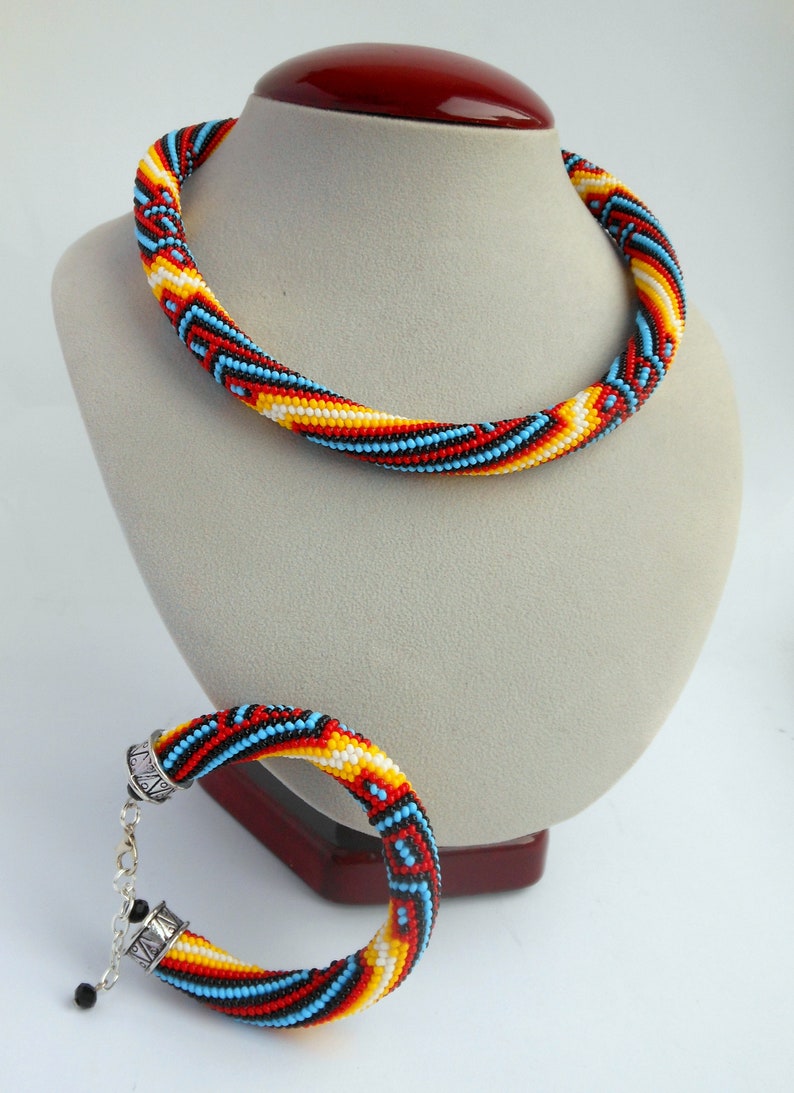 Op de afbeelding: Een kralenketting en armband set. De sieraden hebben een spiraalvormig ontwerp met rode, gele, blauwe en zwarte kralen. De ketting wordt op een mannequin getoond en de armband ligt ernaast.