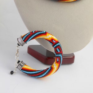 Op de afbeelding: Een kralen sieradenset met een armband en ketting. De armband en ketting zijn gemaakt met kleine, kleurrijke kralen in een spiraalpatroon van rood, zwart, geel en blauw. De armband heeft een zilveren sluiting.