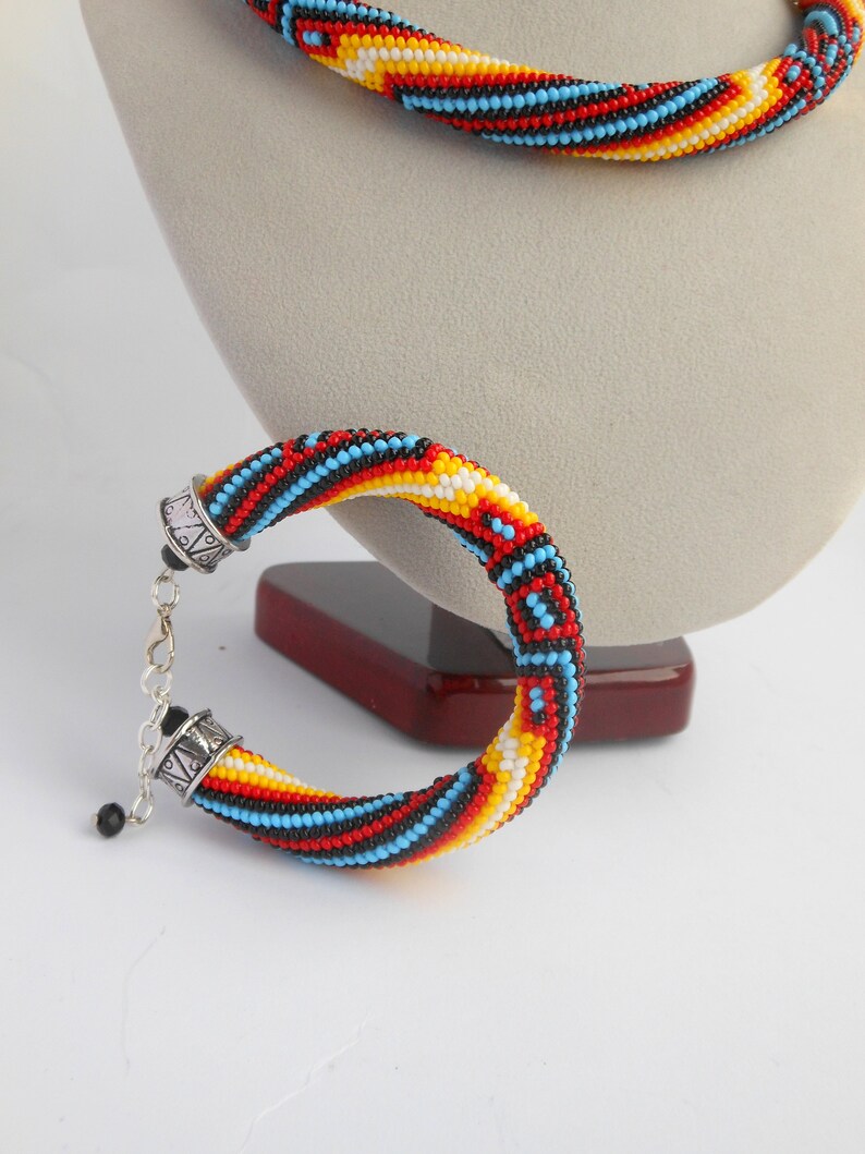 Op de afbeelding: Een kralen sieradenset met een armband en ketting. De armband en ketting zijn gemaakt met kleine, kleurrijke kralen in een spiraalpatroon van rood, zwart, geel en blauw. De armband heeft een zilveren sluiting.