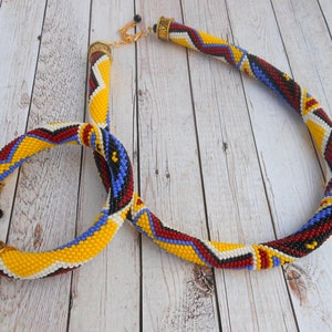 May include: A beaded necklace and bracelet set with a geometric pattern in yellow, red, blue, black, and white. The necklace has a gold clasp and the bracelet has a gold toggle clasp.