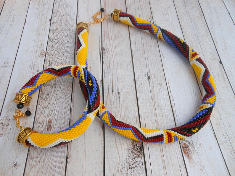 May include: A beaded necklace and bracelet set with a geometric pattern in yellow, red, blue, black, and white. The necklace has a gold clasp and the bracelet has a gold toggle clasp.