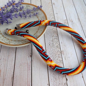 Op de afbeelding: Een kralenketting met een kleurrijk geometrisch patroon. De ketting heeft rode, gele, zwarte en blauwe kralen. De uiteinden hebben zilverkleurige metalen doppen. De ketting ligt op een witte plaat met een gouden rand, met lavendel.