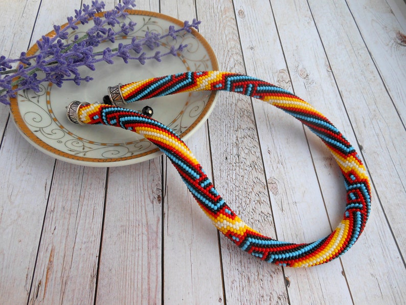Op de afbeelding: Een kralenketting met een kleurrijk geometrisch patroon. De ketting heeft rode, gele, zwarte en blauwe kralen. De uiteinden hebben zilverkleurige metalen doppen. De ketting ligt op een witte plaat met een gouden rand, met lavendel.