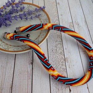 Op de afbeelding: Een kleurrijke kralenketting met een geometrisch patroon in rood, geel, zwart en blauw. De ketting ligt op een wit bord met een gouden rand, naast lavendel.