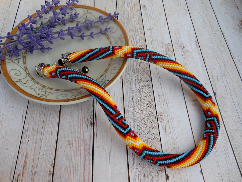 Op de afbeelding: Een kleurrijke kralenketting met een geometrisch patroon in rood, geel, zwart en blauw. De ketting ligt op een wit bord met een gouden rand, naast lavendel.