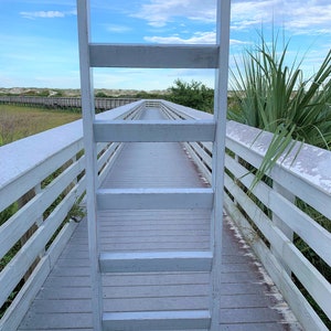 May include: A white wooden ladder leaning against a white wooden boardwalk. The boardwalk is surrounded by green grass and trees. The sky is blue with white clouds.
