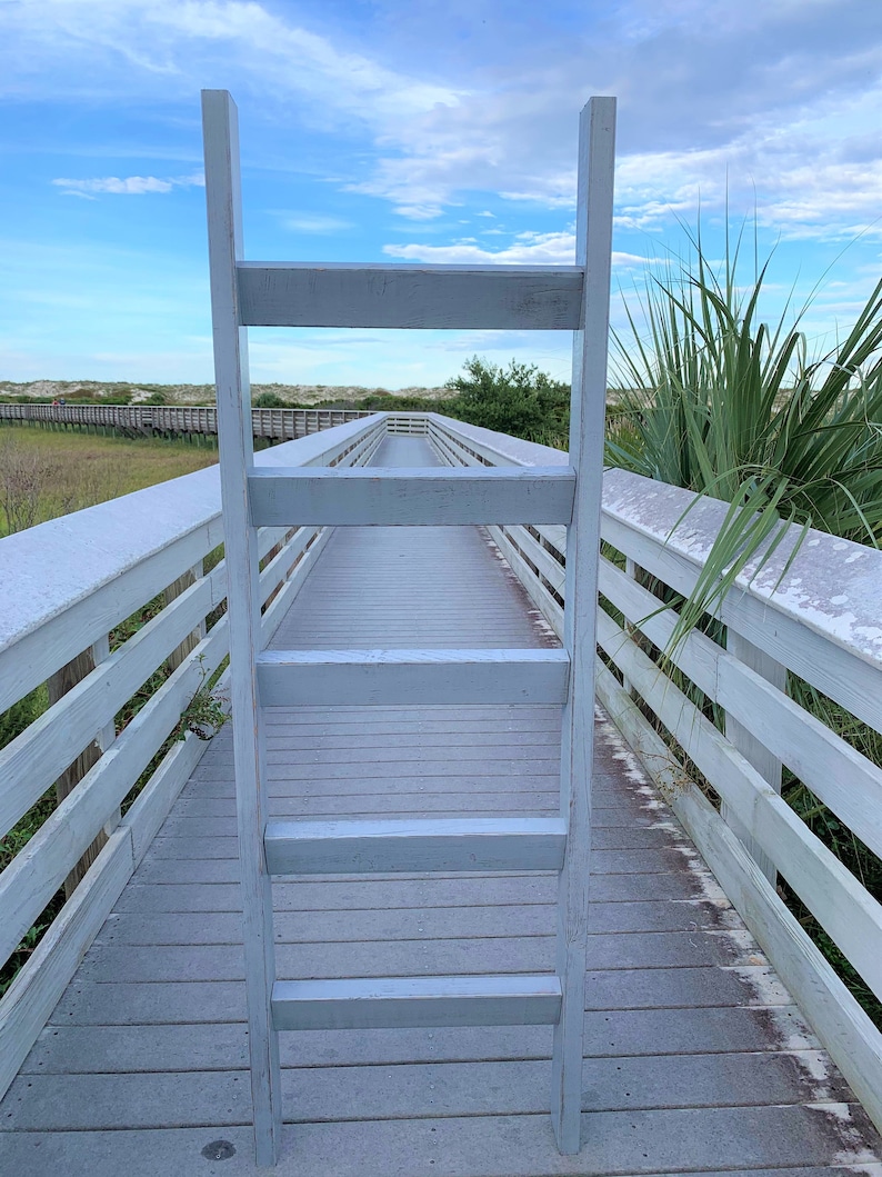 May include: A white wooden ladder leaning against a white wooden boardwalk. The boardwalk is surrounded by green grass and trees. The sky is blue with white clouds.