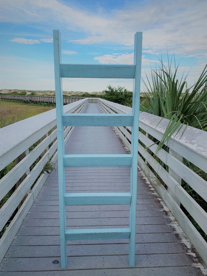 May include: A light blue wooden ladder leaning against a white wooden railing on a boardwalk. The boardwalk is made of wood planks and is surrounded by green grass and sand.