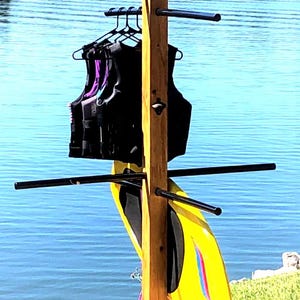May include: Wooden watersports gear rack with black horizontal bars. The rack holds black life vests and a yellow wakeboard. The background features a lake and trees.