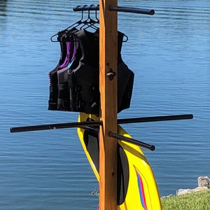 May include: A wooden stand with four legs and multiple black metal bars for hanging water sports equipment. A yellow wakeboard is leaning against the stand. Several life jackets in black, blue, and purple are hanging from the bars. The stand is on a wooden deck overlooking a lake.