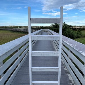 May include: A white wooden ladder leaning against a wooden walkway with railings. The walkway is made of gray wood planks and leads to a sandy beach in the distance. The sky is blue with white clouds.