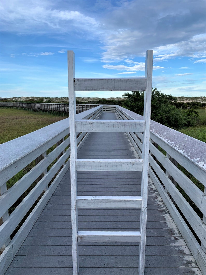 May include: A white wooden ladder leaning against a wooden walkway with railings. The walkway is made of gray wood planks and leads to a sandy beach in the distance. The sky is blue with white clouds.