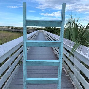 May include: A light blue wooden ladder leaning against a white wooden boardwalk leading to a beach. The boardwalk is surrounded by green grass and sand dunes.