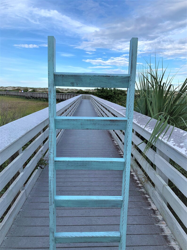 May include: A light blue wooden ladder leaning against a white wooden boardwalk leading to a beach. The boardwalk is surrounded by green grass and sand dunes.
