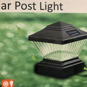 May include: A black solar post light with a clear glass top and a solar panel on the top. The light is sitting on a green grassy surface. The text "Solar Post Light" is visible on the packaging.