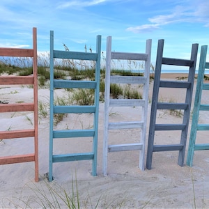 May include: Five wooden ladders painted in different colors: coral, teal, white, gray, and light blue. The ladders are standing upright in a row on a sandy beach.