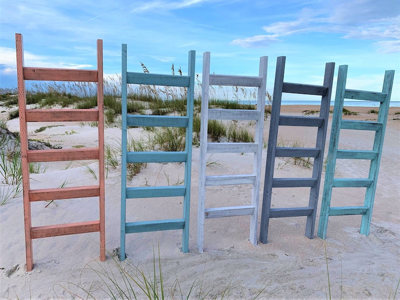 May include: Five wooden ladders painted in different colors: coral, teal, white, gray, and light blue. The ladders are standing upright in a row on a sandy beach.