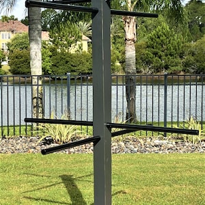 May include: Black wooden towel rack with six arms, standing on a cross-shaped base. The rack is in a backyard setting with a fence and a pond in the background.