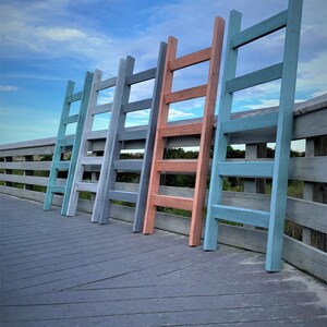 May include: Five wooden step ladders painted in different colors, including blue, gray, and coral, are lined up on a wooden deck. The ladders are all the same size and have four steps.