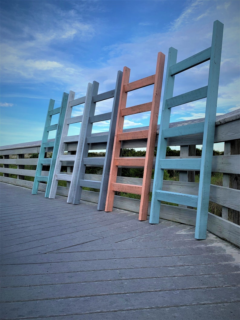 May include: Five wooden step ladders painted in different colors, including blue, gray, and coral, are lined up on a wooden deck. The ladders are all the same size and have four steps.