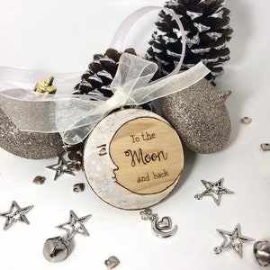 May include: A wooden moon-shaped ornament with the text "To the Moon and back". The ornament has a white painted border and a face design. The ornament is hanging with a white ribbon. Decorative silver stars, bells, and a glittery acorn are scattered around.