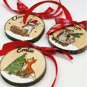 May include: Three wooden Christmas ornaments with red ribbons. Each ornament features a hand-painted winter scene with a name: Claire, Madelyn, and Emilie. The Claire ornament shows a deer wearing a Santa hat. The Madelyn ornament shows a mouse. The Emilie ornament shows a fox.