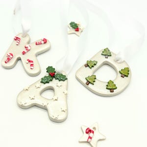 May include: Christmas-themed ornaments in the shape of the letters K, A, and D, along with a star. The ornaments are off-white with festive decorations like candy canes, Christmas trees, and holly. Each ornament has a white ribbon for hanging.