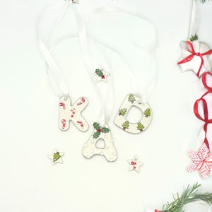 May include: White ceramic Christmas ornaments spelling "KAD" with candy cane and Christmas tree designs. Each letter is strung with a white ribbon for hanging. Additional star-shaped ornaments with holly and tree designs are also present.