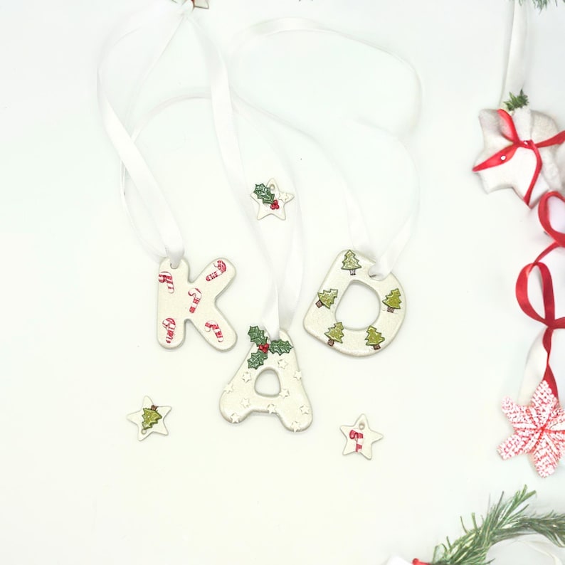 May include: White ceramic Christmas ornaments spelling "KAD" with candy cane and Christmas tree designs. Each letter is strung with a white ribbon for hanging. Additional star-shaped ornaments with holly and tree designs are also present.