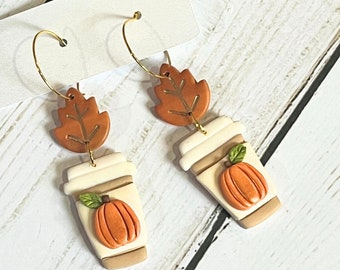 May include: A pair of fall-themed earrings. Each earring features a gold hoop, a brown leaf, and a coffee cup with a pumpkin. The coffee cup is cream-colored with a brown base and a pumpkin with a green leaf.