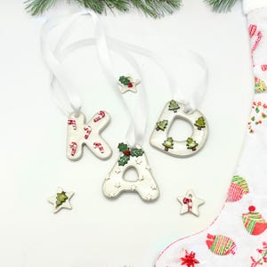 May include: White ceramic Christmas ornaments spelling "KAD" with red and white candy cane and green Christmas tree designs. Each letter is strung with a white ribbon. Additional star-shaped ornaments with similar designs are also present.