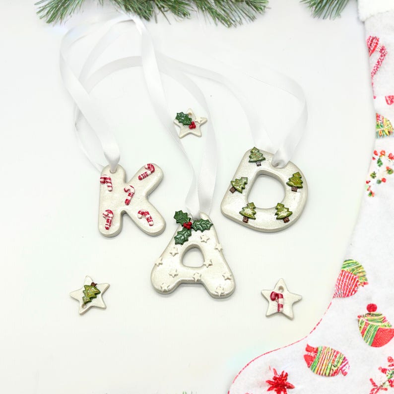 May include: White ceramic Christmas ornaments spelling "KAD" with red and white candy cane and green Christmas tree designs. Each letter is strung with a white ribbon. Additional star-shaped ornaments with similar designs are also present.